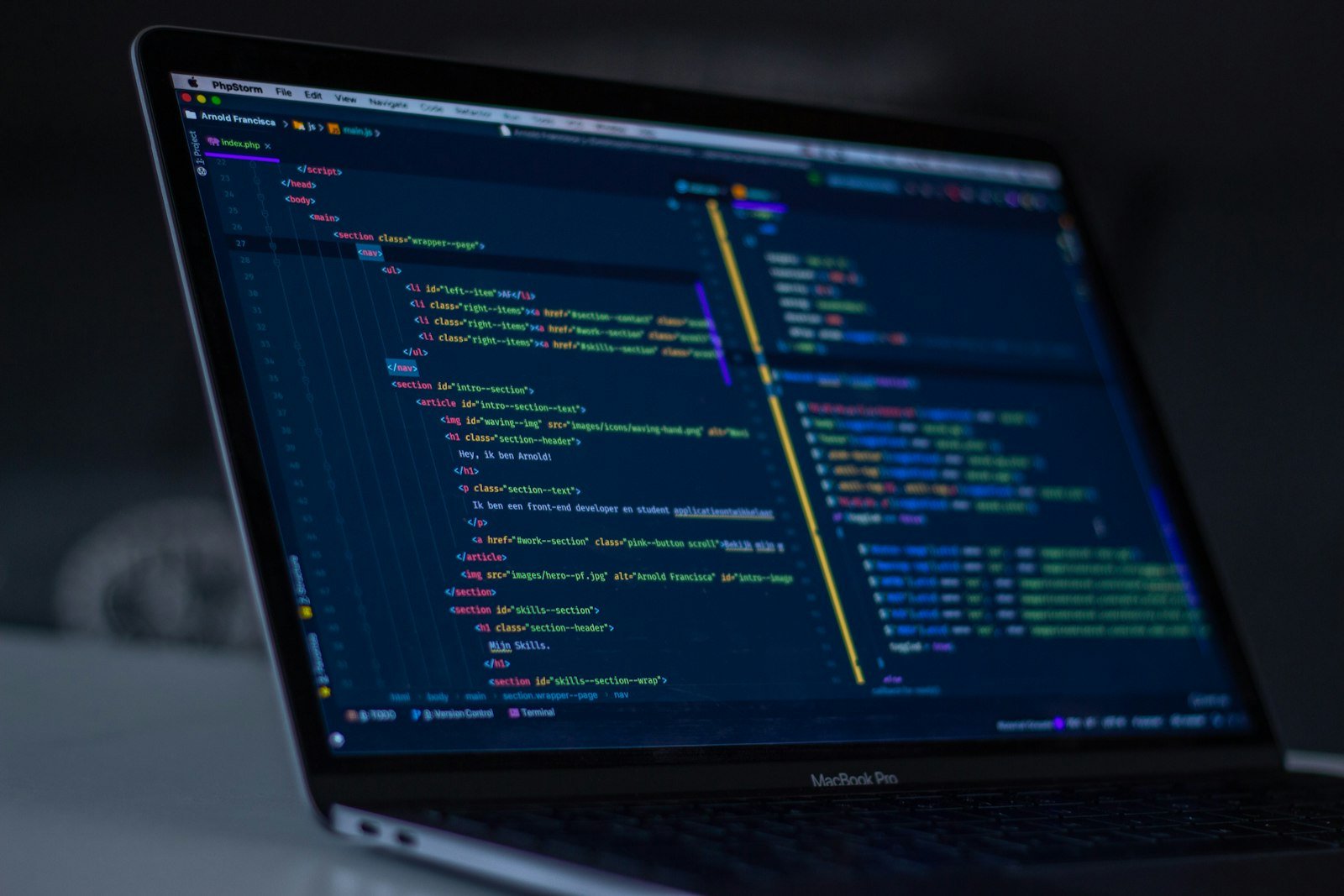Portátil numa sala pouco iluminada, com código HTML aberto no editor e um painel de ficheiros à direita.